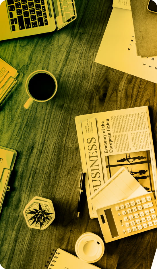 An aerial shot of a busy desk at a digital agency with items such as a laptop, notebook, newspaper and calculator on.